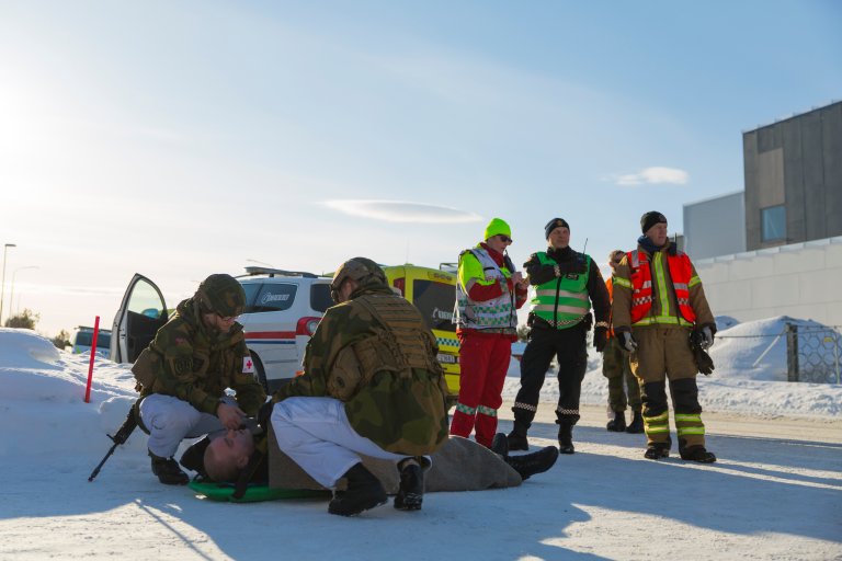 To soldater bøyer seg ned over mann som ligger på båre. I bakgrunnen står ulike nødetater