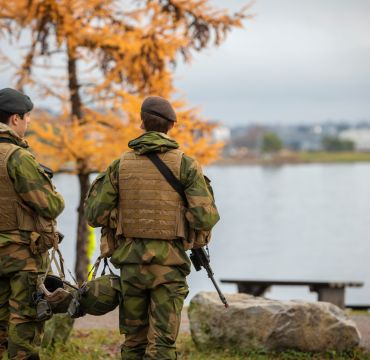 to soldater står med ryggen til og ser utover et vann. Det er høstfarger på trærne. 