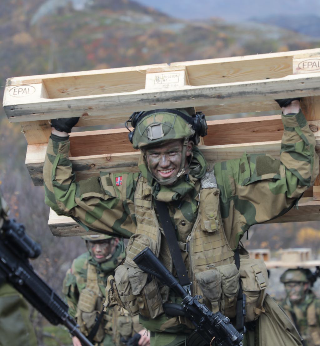 Soldat med full utrustning og kamuflasjemaling i ansiktet løper mens han bærer en europall på ryggen.