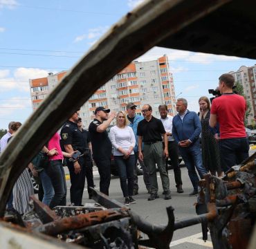 En rekke personer står foran et ødelagt kjøretøy. En politimann snakker til forsamlingen.