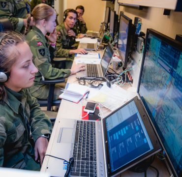 en ung kvinne i grønn militæruniform og norsk flagg på armen sitter med hodetelefoner og ser på flere dataskjermer