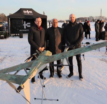Tre menn poserer bak en drone på en snølagt flyplass.