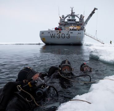 Tre dykkere i havet, ved iskanten. I bakgrunnen ser vi kystvaktens isbryter. 