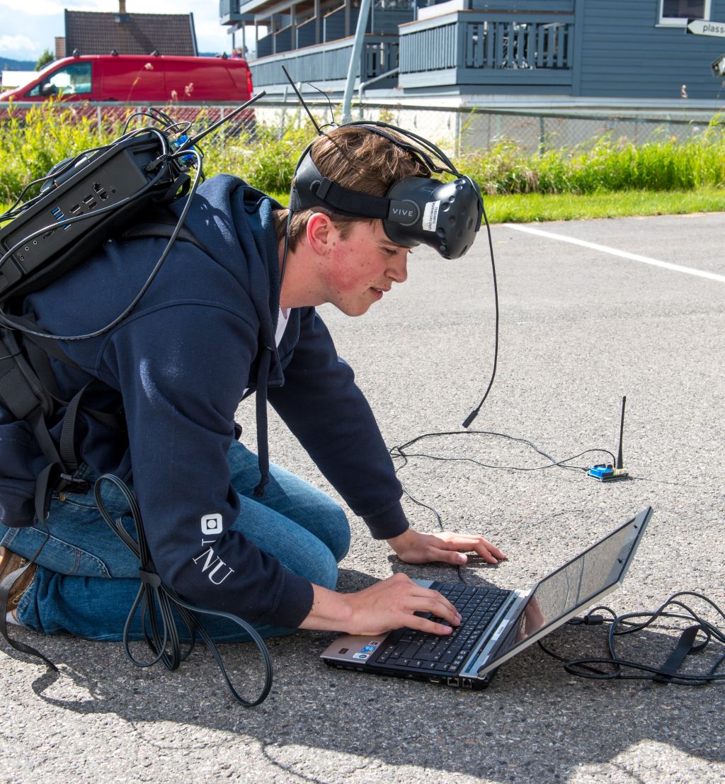 sommerstudent sitter ute med en laptop på bakken og utstyr i en sekk og på hodet