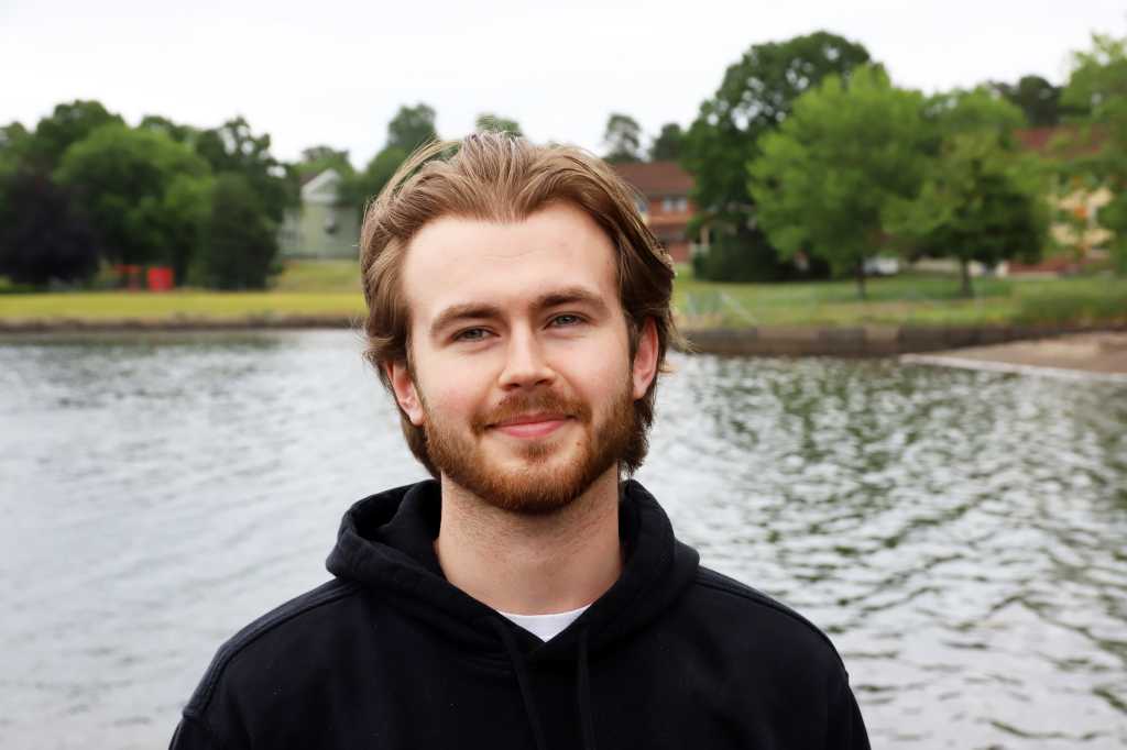 Ung mann i hettegenser står utendørs med havet i bakgrunnen. Ser rett i kamera og smiler