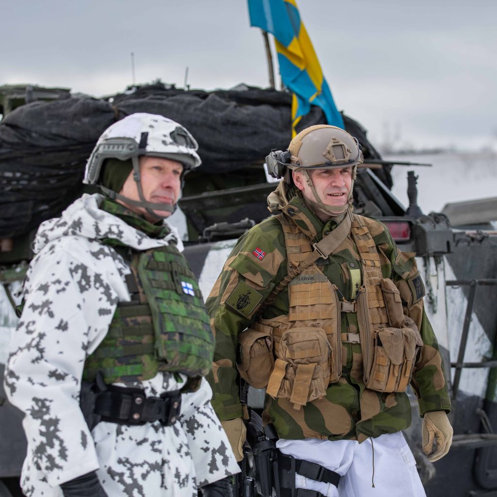 En finsk og en norsk soldat står i vinterbekledning ute foran et kjøretøy med svensk flagg