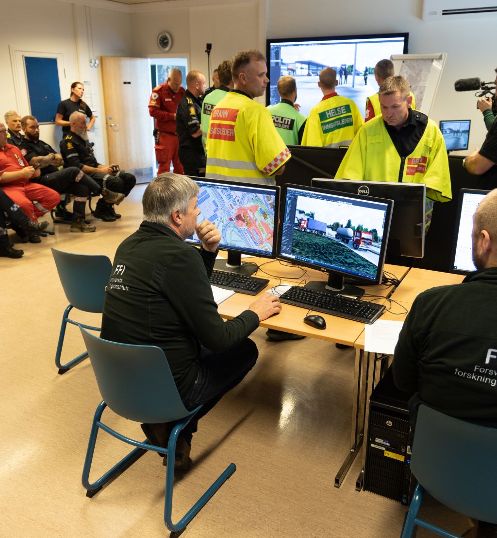 Politi, brann og ambulansepersonell står i et klasserom og diskuterer hvordan de skal løse en krisesituasjon der en tankbil full av amoniakk har kollidert og sprunget lekk. På en skjerm på veggen kan vi se en virtuell verden der krsien utspiller seg. Langs vekggen sitter kursdeltagere på to stolrader og ser på. I forgrunnen ser vi to FFI-ansatte som styrer spillet.