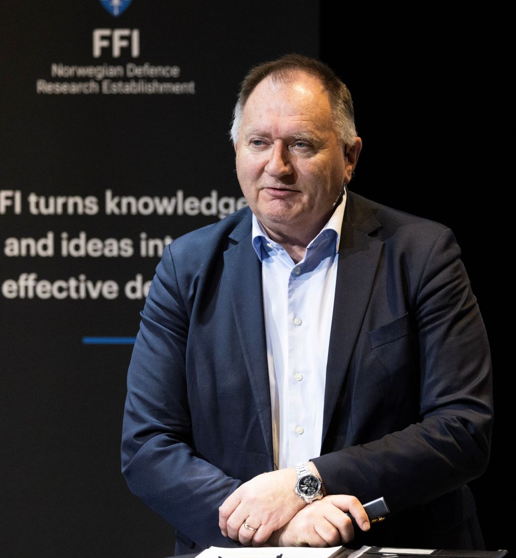 eldre mann står bak et høyt bord og snakker henvendt til et publikum. 