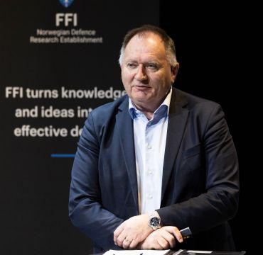 eldre mann står bak et høyt bord og snakker henvendt til et publikum.