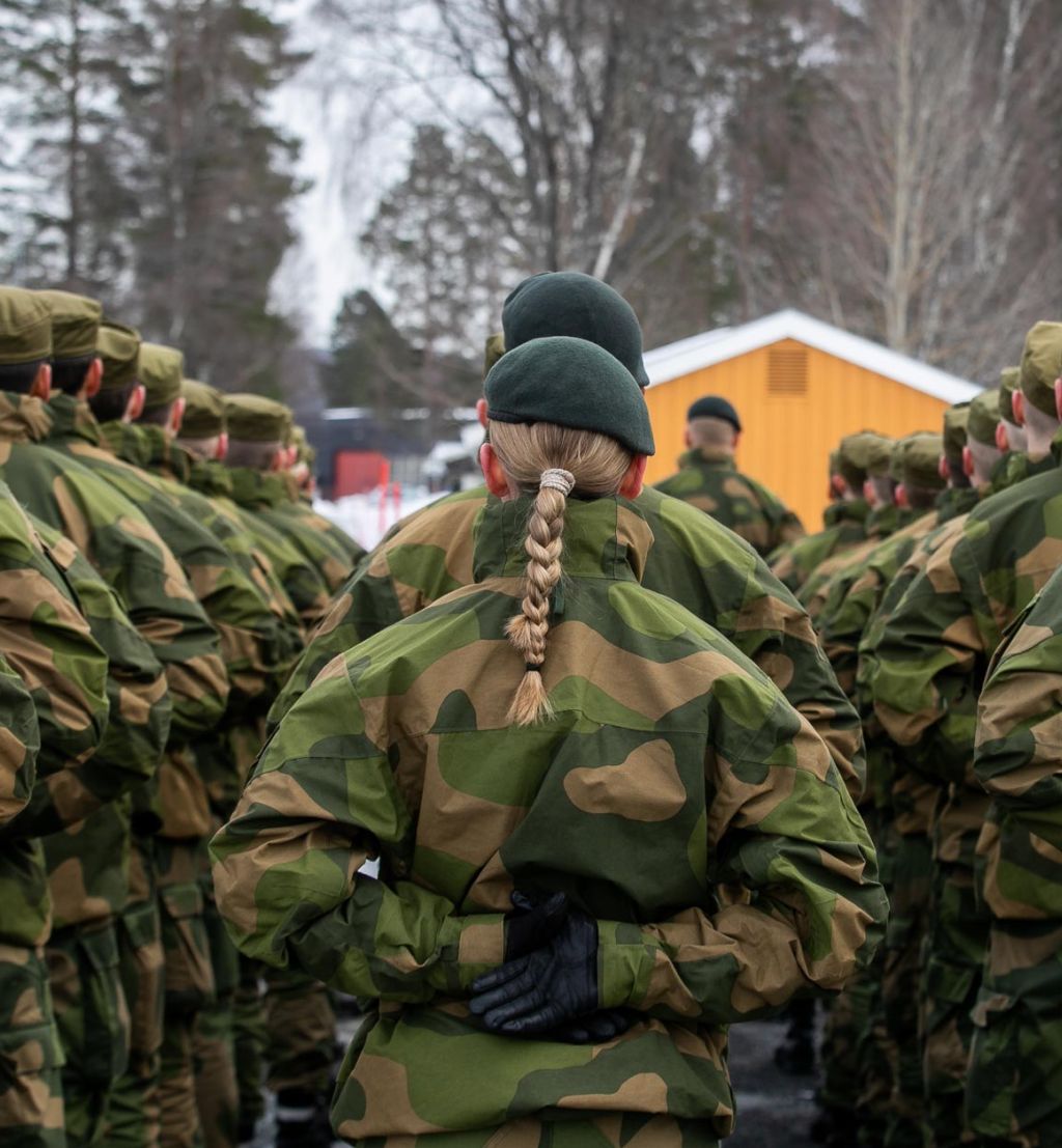 Soldater på oppstilling i grønn feltuniform. Alle står med ryggen til. I midten en kvinne med en lang flette.