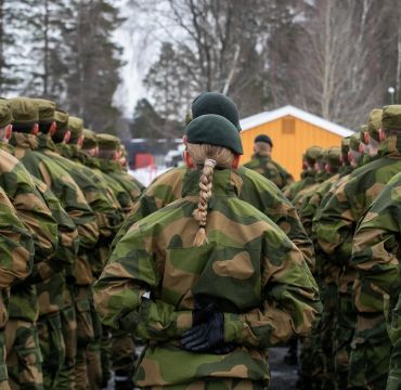 Soldater på oppstilling i grønn feltuniform. Alle står med ryggen til. I midten en kvinne med en lang flette.