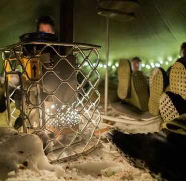 Vi er inne i et telt. Det er nærbilde av en ovn og flere soldatføtter som varmer seg på den