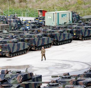 et oversiktsbilde viser en stor plass med mange stridsvogner og andre militære kjøretøy som er parkert. Midt på plassen går en mann i hæruniform