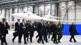 Menn i dress går rundt i en stor hangar med et moderne hvitt jagerfly i bakgrunnen