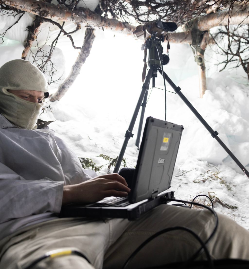 Soldat sitter i snøhule med en liten datamskin foran seg. Han har buff foran ansiktet. I åpningen står en kikkert på stativ.
