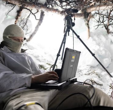 Soldat sitter i snøhule med en liten datamskin foran seg. Han har buff foran ansiktet. I åpningen står en kikkert på stativ.