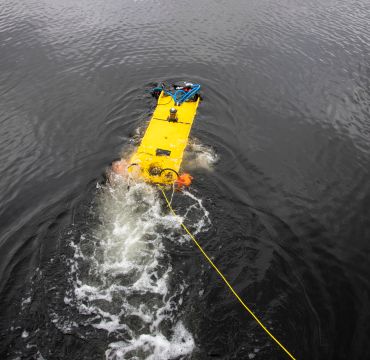 En liten, gul undervannsfarkost kjører framover i vannoverflaten med en gul kabel etter seg.