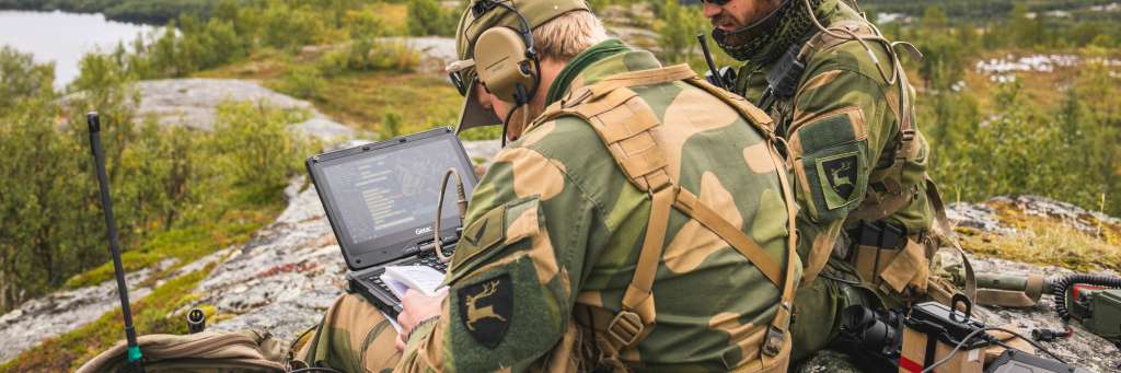 To militærkledde sitter med PC og radioutstyr på en fjellknaus i høstlandskap